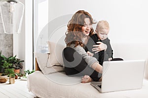 Young cheerful woman sitting on sofa with her little handsome son happily using laptop together. Mom with baby boy