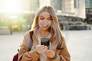 Young cheerful woman using cell phone and texting message on city street. Happy girl holding smart phone in hands and smiling.