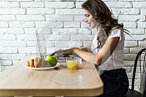 Young cheerful woman using on laptop computer and eating fruits in the kitchen in the morning