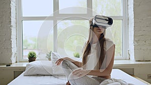 Young cheerful woman wearing virtual reality headset playing 360 VR video game sitting in the bed at home