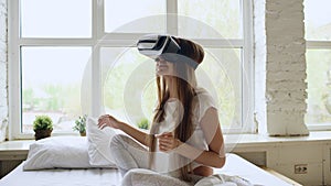 Young cheerful woman wearing virtual reality headset watching 360 VR video movie sitting in the bed at home