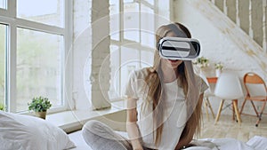 Young cheerful woman wearing virtual reality headset watching 360 VR video movie sitting in the bed at home