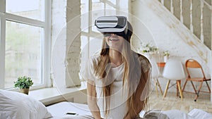 Young cheerful woman wearing virtual reality headset watching 360 VR video movie sitting in the bed at home