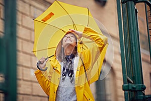 A young cheerful woman with a yellow raincoat and umbrella who is enjoying while listening to the music and walking the city on a