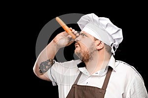 Young chef in hat and apron holding raw carrot near nose and looking away