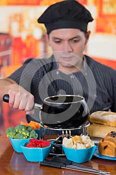 Young cheff preparing a gourmet Swiss fondue dinner with assorted cheeses and a heated pot of cheese fondue and some