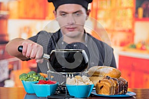 Young cheff preparing a gourmet Swiss fondue dinner with assorted cheeses and a heated pot of cheese fondue and some