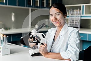 Young chemist in white coat working with microscope and smiling at camera in lab