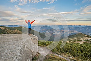 Young child boy hiker standing with raised hands in mountains enjoying view of amazing mountain landscape at sunset