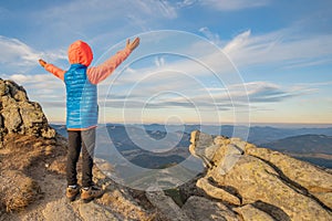 Young child boy hiker standing with raised hands in mountains enjoying view of amazing mountain landscape at sunset