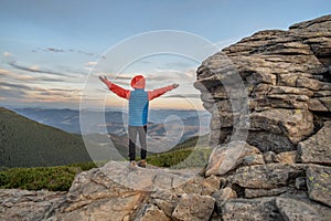 Young child boy hiker standing with raised hands in mountains enjoying view of amazing mountain landscape at sunset