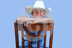 Young child cowboy wears blue plaid shirt and white cowboy hats sits on chair with arms crossed, smiling at camera, studio, close