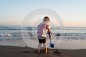 A young child gazes into the sea while holding a blue shovel on the beach, with the warm glow of sunset lighting the