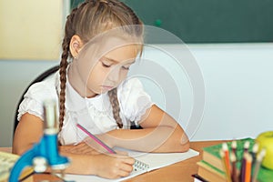 Young child girl drawing or writing with colorful pencils in notebook in school over blackboard