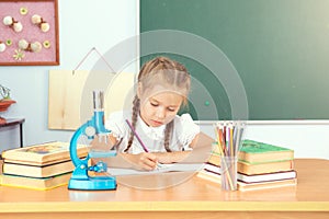 Young child girl drawing or writing with colorful pencils in notebook in school over blackboard