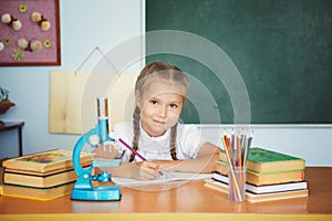 Young child girl drawing or writing with colorful pencils in notebook in school over blackboard