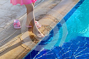 Young child girl in pink dress and shoes standing barefoot at the edge of a swimming pool try with her leg cool blue water,