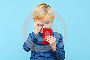 Young child holding smartphone, watching the screen, rubbing his eyes, isolated over pastel blue background.