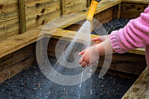 Young child holds hands under water hose spray at vegetable box in garden.