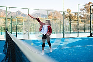 A young child in a red jacket hides behind a vibrant orange padel racket, with the court and players blurred in the