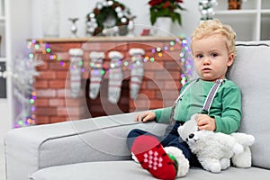 A young child sits on a gray sofa against the backdrop of a fuzzy brick fireplace