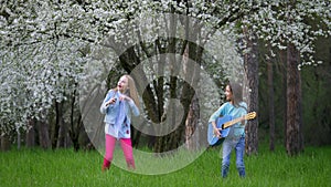 Young children girlfriends have fun dancing singing and playing blue acoustic guitar springtime blooming tree into the park. conce