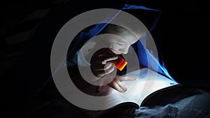 Young children reading a book hidden under the bed covers