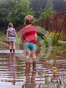 Young children in rubber boots pull toys on a rope along a country sandy road with puddles after rain