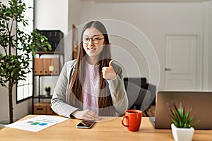 Young chinese business worker wearing business style sitting on desk at office smiling happy and positive, thumb up doing