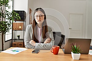 Young chinese business worker wearing business style sitting on desk at office skeptic and nervous, disapproving expression on photo