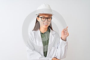 Young chinese engineer woman wearing coat helmet glasses over isolated white background with a big smile on face, pointing with