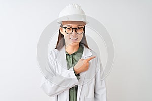 Young chinese engineer woman wearing coat helmet glasses over isolated white background cheerful with a smile of face pointing