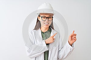 Young chinese engineer woman wearing coat helmet glasses over isolated white background smiling and looking at the camera pointing