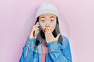 Young chinese girl having conversation talking on the smartphone covering mouth with hand, shocked and afraid for mistake