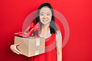 Young chinese girl holding red high heel shoes and package looking positive and happy standing and smiling with a confident smile
