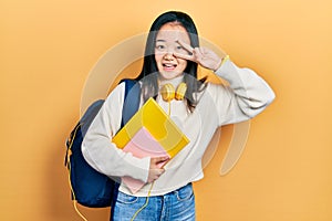 Young chinese girl holding student backpack and books doing peace symbol with fingers over face, smiling cheerful showing victory