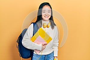 Young chinese girl holding student backpack and books with a happy and cool smile on face