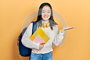 Young chinese girl holding student backpack and books smiling cheerful presenting and pointing with palm of hand looking at the