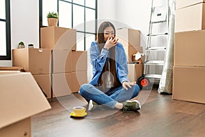 Young chinese girl sitting on the floor at new home smelling something stinky and disgusting, intolerable smell, holding breath