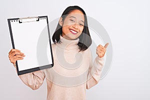 Young chinese inspector woman holding clipboard standing over isolated white background happy with big smile doing ok sign, thumb