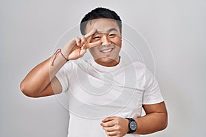 Young chinese man standing over white background doing peace symbol with fingers over face, smiling cheerful showing victory