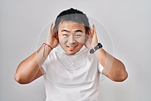 Young chinese man standing over white background trying to hear both hands on ear gesture, curious for gossip