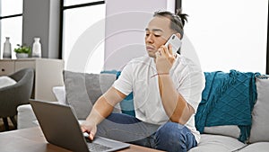 Young chinese man using laptop talking on smartphone sitting on sofa at home