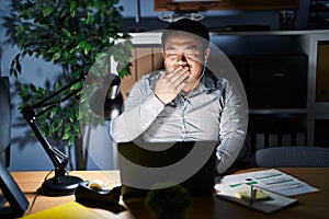 Young chinese man working using computer laptop at night bored yawning tired covering mouth with hand