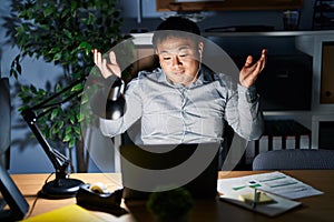 Young chinese man working using computer laptop at night clueless and confused expression with arms and hands raised