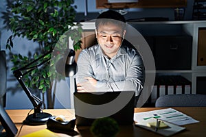 Young chinese man working using computer laptop at night happy face smiling with crossed arms looking at the camera