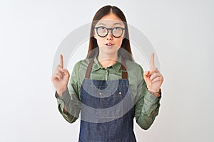 Young chinese shopkeeper woman wearing apron and glasses over isolated white background amazed and surprised looking up and