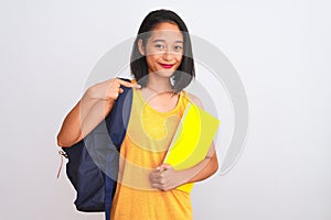 Young chinese student woman wearing backpack holding book over isolated white background with surprise face pointing finger to