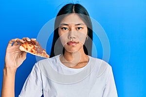Young chinese woman eating tasty pepperoni pizza thinking attitude and sober expression looking self confident