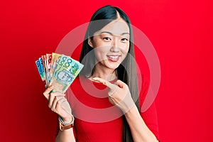 Young chinese woman holding australian dollars smiling happy pointing with hand and finger